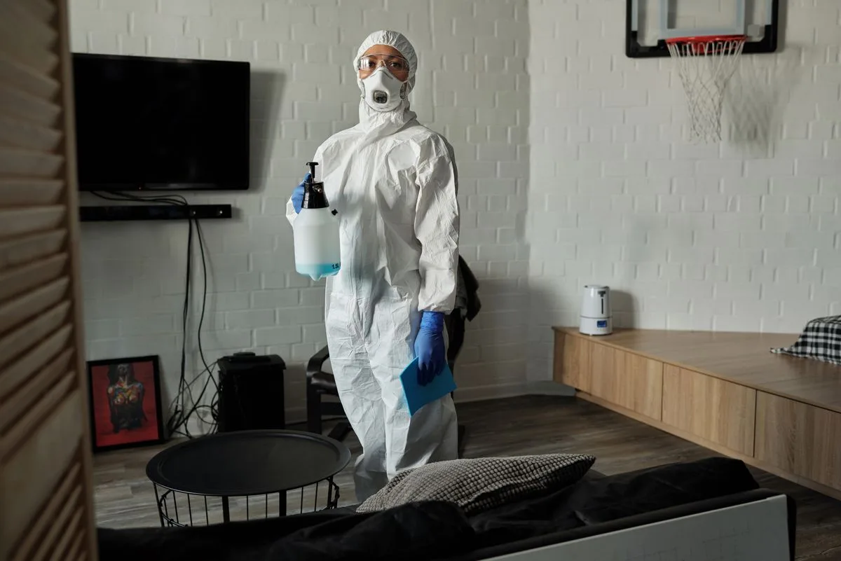 Technician in full protective equipment with a pump sprayer standing in a living room